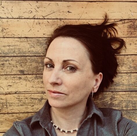 Headshot of Shelley Senter. She wears a gray button up and a beaded necklace. Her hair is dark brown and and put up in an updo. She looks into the camera with a neutral expression. Photo courtesy of the artist.
