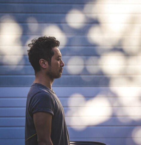 Photo of Takahiro in front of a slated wall with shadows of a tree. He is standing profile in a grey shirt.