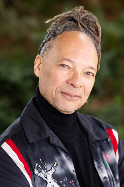 A person wearing a black turtleneck shirt sits for a portrait, and tilts his head with green eyes towards the viewer, his head crowned by reddish dreadlocks and a 'mona lisa' smile. Photo courtesy the artist.