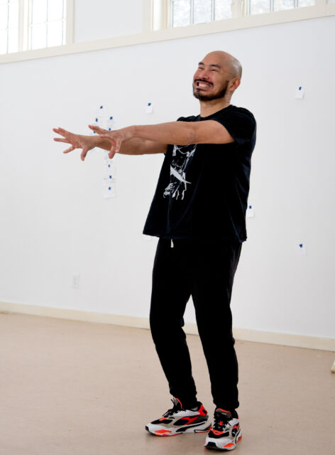 Justin stands in a bright, airy room wearing a black graphic T-shirt, jeans, and sneakers with red accents. His arms extend in front of his body with fingers reaching forward. He is grinning ear-to-ear. Photo courtesy of the artist.