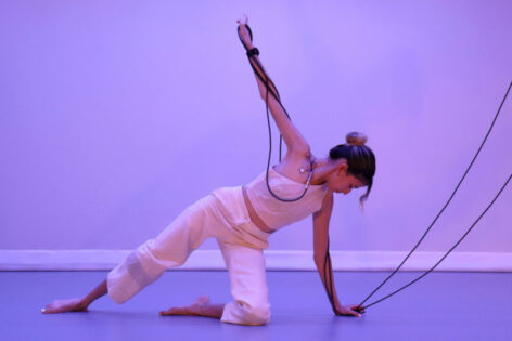 Crouching on one knee with one arm up reaching towards the ceiling, wrapped by a tangle of audio cables, Yo-Yo performs with contact microphones attached to her shoulder and back. She is a thin, Taiwanese American femme, her hair in a bun with wavy bangs hanging over her face. Her stance is solid and her face is serene. Photo by Buffy.