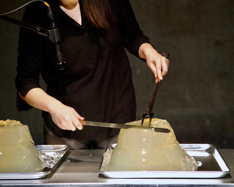 A woman holding a fork and knife, cutting a large jelly. Alison Kuo, The New Joys of Gellies, 2019, Abrons Arts Center. Programmed in conjunction with Extremely absorbent and increasingly hollow, Cuchifritos Gallery + Project Space, New York. Photo by Georgette Maniatis