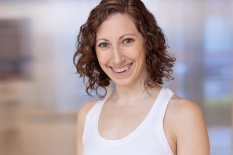 Allie is kneeling on the floor, left leg bent and extended toward the camera. Her toes are flexed off the floor and she is barefoot wearing a white top and green pants. Allie is in the background looking past her foot into the foreground of a light wood floor. Photo courtesy of the artist.