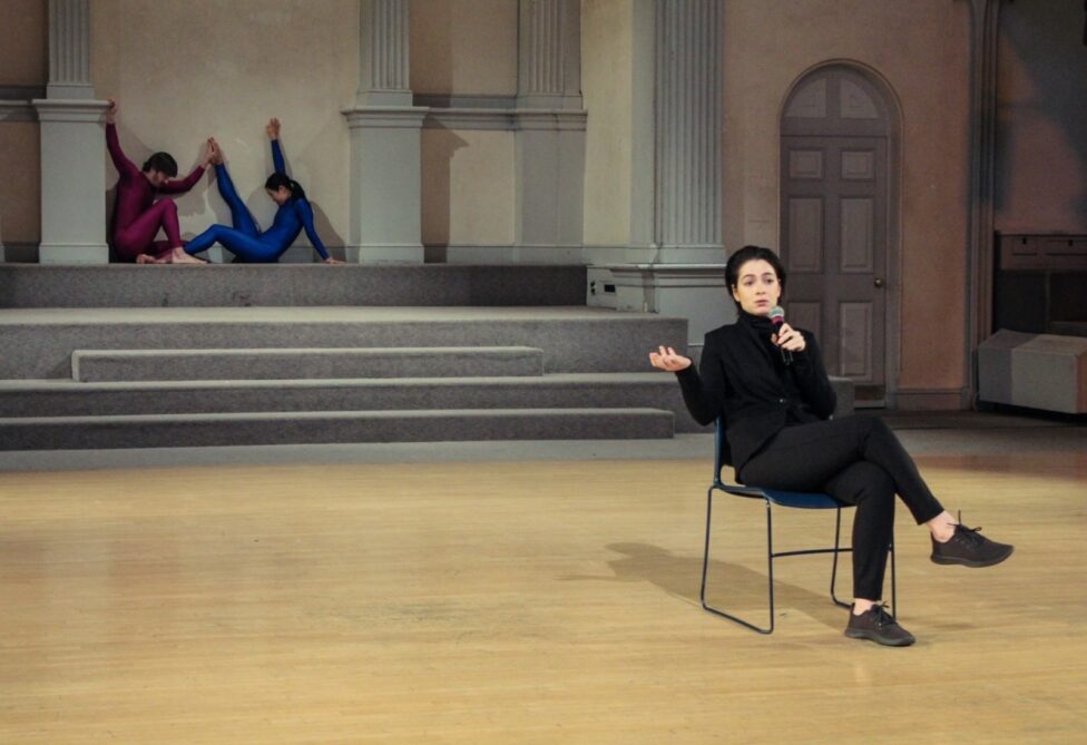 Person sitting in a chair, speaking into a microphone with two dancers moving behind them. Justin Faircloth, Annie Heath, and Mariam Dingilian in Ruth at Danspace, photo by Jessie Young