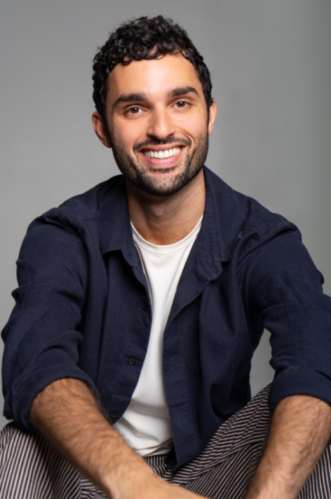 Portrait of Marc Crousillat. He is smiling into the camera sitting with his arms over his knees. He wears a white T-shirt and a dark blue button down top. Photo courtesy of the artist.