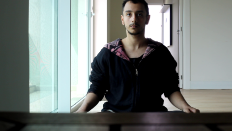 Habib can be seen from the waist up and is wearing a black jacket with a pink geometric hood. In the background, you can see an open door and a white wall. On the side, there is a glass door. Their face has a small amount of short beard, dark brown eyes and hair, light skin, black frame glasses and gold ring piercings in both ears. Photo courtesy of the artist.