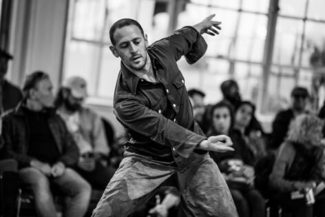 Photo of Jesse Zarrit performing in front of an audience. He stands with his legs wide apart. His torso is leanning forward with one arm reaching across his chest and the other reaching out behind him. His gaze is cast downward. Photo By Robbie Sweeney.