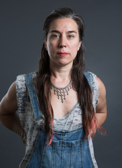 Portrait from waist up of middle age light skinned luciana achugar, femme with long hair below breasts with dyed red ends and brown hair with some grey hairs on top, half pony tail. luciana's arms are bent, hands hidden resting in back of waist, elbows pointing back. eyes looking straight at the camera with red lipstick stained lips; Some wrinkles around mouth and between eyebrows. Photo by Scott Shaw.