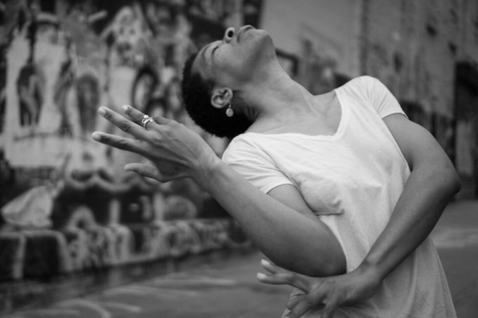 Black and white image of a dancer in a white v-neck t-shirt, leaning backwards and extending a hand. 