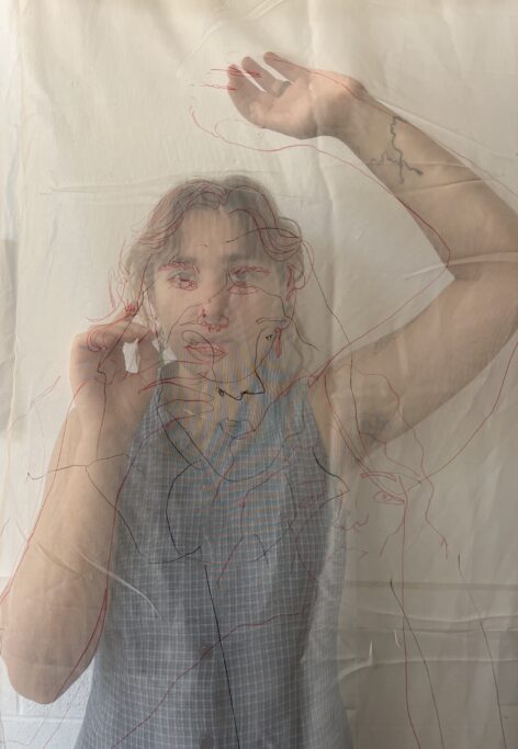 A photo of Jenna behind a transparent fabric with continuous artistic outlines of their figure and face in red and black ink. Their right arm is uo towards their face touching the screen and their left arm is reaching up towards the sky. They are wearing a blue sleeveless gingham collared shirt. Their hair is parted in the middle and falling softly on their forehead. Photo courtesy of the artist.