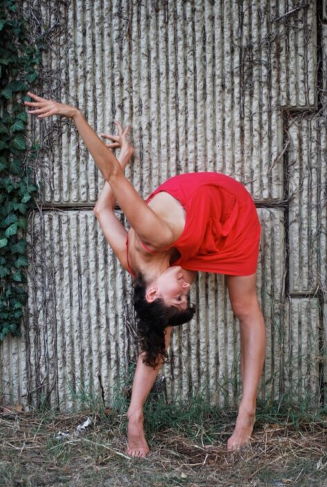 Gabrielle Revlock in a red dress against a grey wall bending forward with her arms up. Photo courtesy of the artist.
