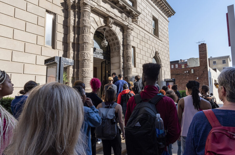 Colorful photo of a crowd gathering outside of a beige building. 