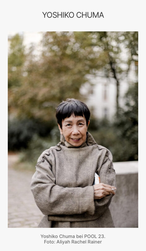 A grinning Yoshiko Chuma wearing a gray tunic and holding a cigarette, standing against a background of blurred trees. Photo by Aliyah Rachel Rainer.