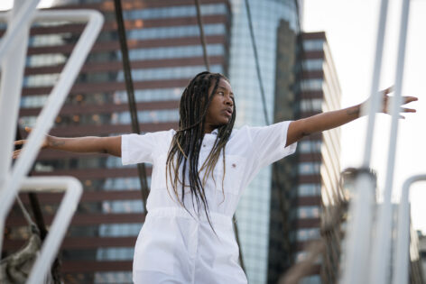 A brown skinned performer with long brown braids in a white jumpsuit. The performer's arms are horizontally outstretched and they are looking intently to the left while they stand on the bow of a ship. A high rise building with reflective windows is in the background. Photo by Cherylynn Tsushima.