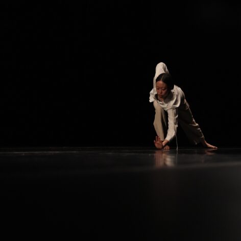 On a darkened stage Clara wears loose beige clothing while draping her torso laterally over her bent right leg as her left leg extends straight behind her. Her right arm forms an angular shape tucked behind her right side body and her gaze is fixed on her left hand’s fingertips. Photo credit to Derek Fowles.