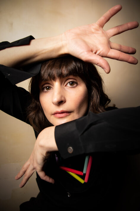 A headshot of Elisabeth Motley, a white disabled woman wearing a black button-up shirt and colorful necklace. She has dark brown hair, her arms rame her head, and her fingers are splayed.