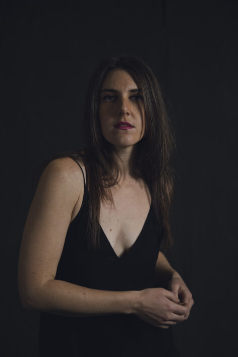 Headshot of Heather (female, long brown hair, green eyes), looking at the camera with a pensive expression, hands softly at one side of waist. She wears a v-neck black shirt and stands against a dark background.