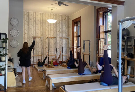 A woman wearing a black sweatshirt and black shorts faces away from the camera holding her right arm up towards the ceiling in a gesture designed to inspire a lifting action. Six people lay on tan mats on their back with a blue ball resting underneath their hips. They hold their bent right knee firmly with both hands as their left leg is long and straight reaching towards the ceiling with a flexed foot. They are in a Pilates studio with brown wood floor and white walls. A photo by Aline Pilates.