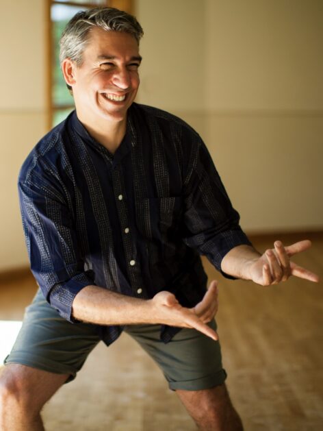Peter, an Italian-American man, stands with his arms outstretched to one side, fingers extended, bending one knee and smiling exuberantly. Photo by Anna M. Maynard.