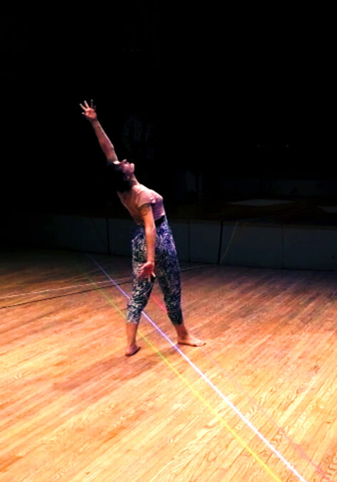 Performance photo of Sarah White-Ayón. A white woman with black hair stands in fourth position with her right foot front and her back facing the audience. She is arching upwards looking towards her left hand that is maximally outstretched above her head while her right hand is outstretched oppositional to her left hand. She stands on a hard wood floor with 3 lines of colored tape, yellow, white and pink attached to the floor passing underneath her feet. She wears black and white speckled pants and a pink t-shirt. The background behind her obscured, almost black. Photo courtesy of the artist.