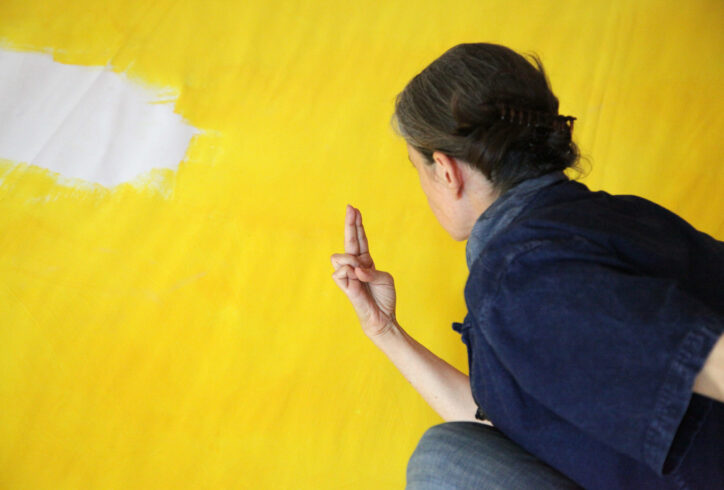 Daria holds her index and middle finger together and looks at it. We see the back of her head, her hair in a low bun while she crouches in front of a bright yellow wall. Photo courtesy of the artist.