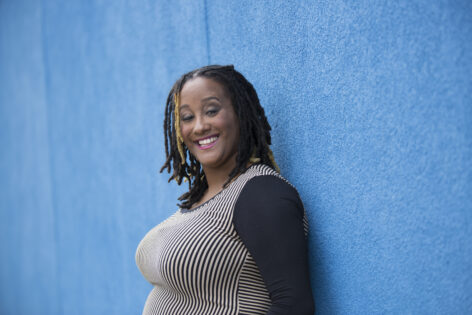 Kayla, a Black voluptuous person, stands with their back against an azure blue concrete wall, and turns their head over their left shoulder to smile joyfully at the camera. They're wearing pink lipstick and their eyebrows are raised in pleasure. Kayla wears a whimsical black and tan thinly-striped shirt with stripes in multiple directions and solid black long sleeves. Their dark locs rest on their shoulders, with some bleached locs throughout their hair. Alison, a white queer femme, lover of lake Michigan, and access coordinator to Circle O, wrote this description from her Chicago home. Photo Credit: Travis Magee.