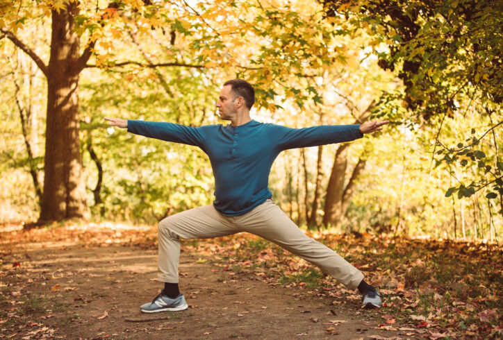 Ori Flomin is in a warrior two position from Yoga practice. He is wearing light beige pants and a blue long sleeve shirt. In this position his right leg is bent to a 90 degree angle while his left leg is fully extended, his spine is vertical and his arms are extended away from each other parallel to the floor. He is standing outside in the park with trees in the background. Photo by Yasmeen Enahora.
