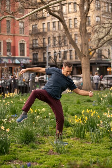 A man dances energetically in a grassy park filled with blooming yellow and white daffodils. He wears a dark blue T-shirt, burgundy pants, and blue sneakers, with one arm bent and the other extended as he lifts one leg in mid-step. Behind him, bare-branched trees and multi-story brick buildings frame the urban setting, with a few pedestrians and cars visible in the background. Photo by Yasmeen Enahora.