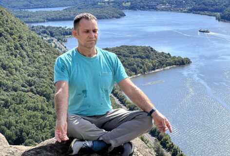 A man sits cross-legged on a rocky cliff, meditating with eyes closed and hands resting on his knees. He wears a turquoise T-shirt, gray pants, and sneakers. Behind him stretches a sweeping view of a winding river bordered by lush green hills, under a bright blue sky dotted with fluffy white clouds. The sunlight reflects off the water, and a boat can be seen traveling along the river. The scene conveys a sense of peace, balance, and connection with nature. Photo by Tony Bordonaro