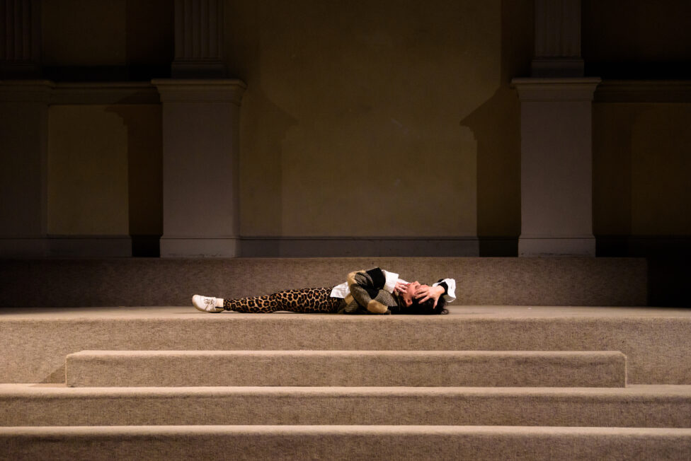 Sam lays on her back at the top of a set of beige stairs, wearing white sneakers, leopard leggings, a white shirt, and a black and gold multi-colored jacket. Her hands touch the top of her head and face.