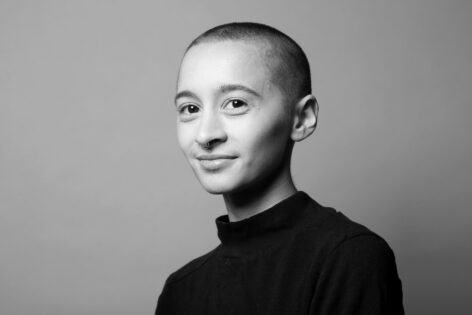 Portrait of Nikaio Bulan Sahar Thomashow. They look into the camera with a small grin. He wears a black turtle neck top and has a shaved head. Photo courtesy of the artist.
