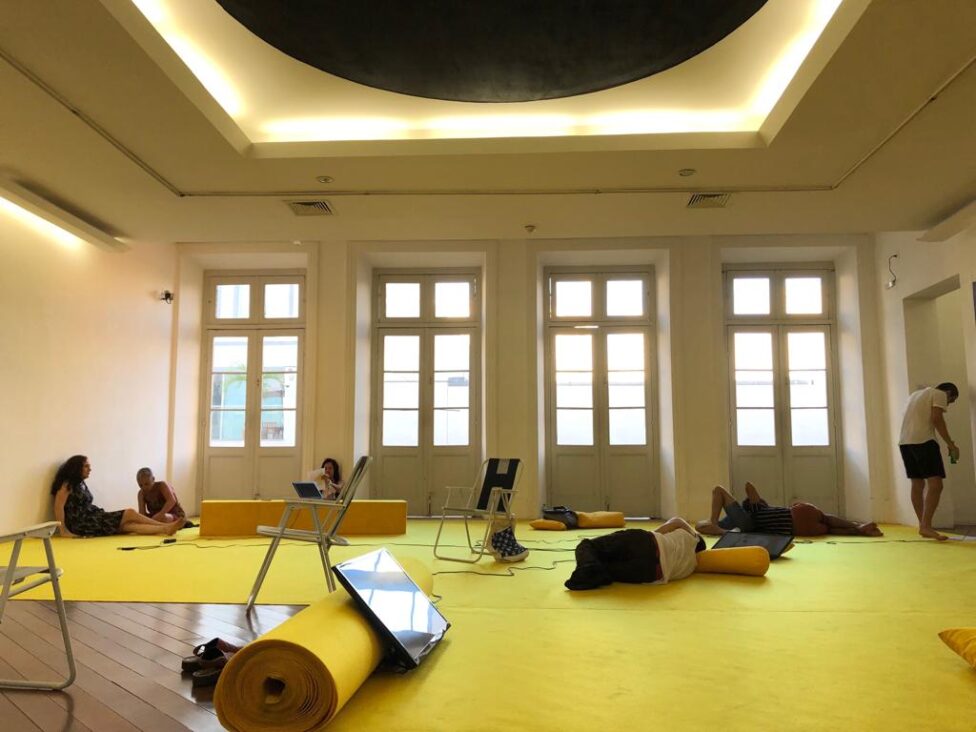 A group of people interact with Sofia's installation, which is occurring in a brightly lit room with yellow carpet, beach chairs, and TV screens scattered intermittently throughout the space. Photo courtesy of the artist.