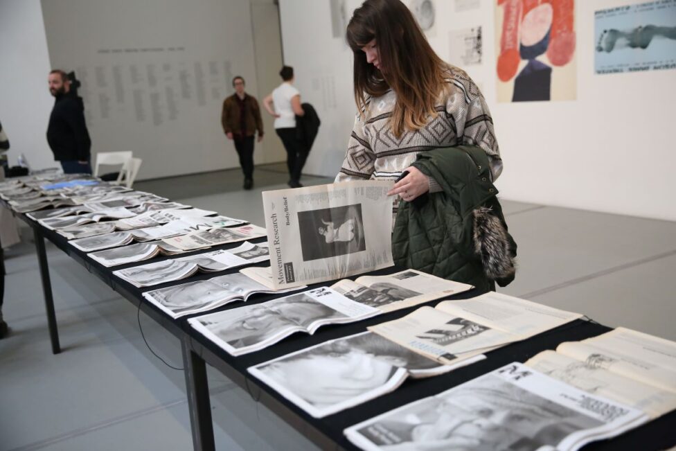 A table at MoMa with different issues of the Movement Research Performance Journal laid out. A person in a patterned sweater with a green coat draped over their arm looks through one. A table at MoMa with different issues of the Movement Research Performance Journal laid out. A person in a patterned sweater with a green coat draped over their arm looks through one. Photo by Paula Court.