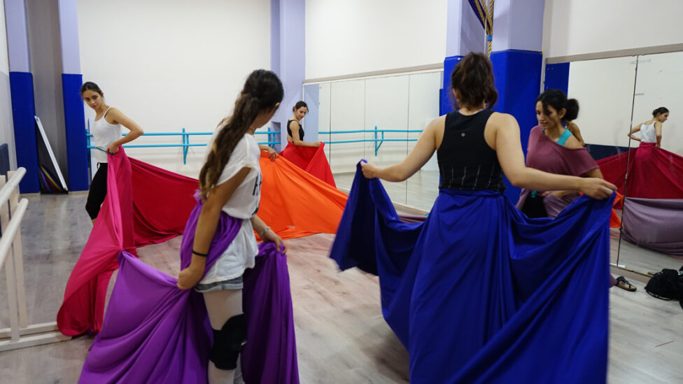 Five or six dancers in a studio, connected by holding one another's brightly colored skirts.