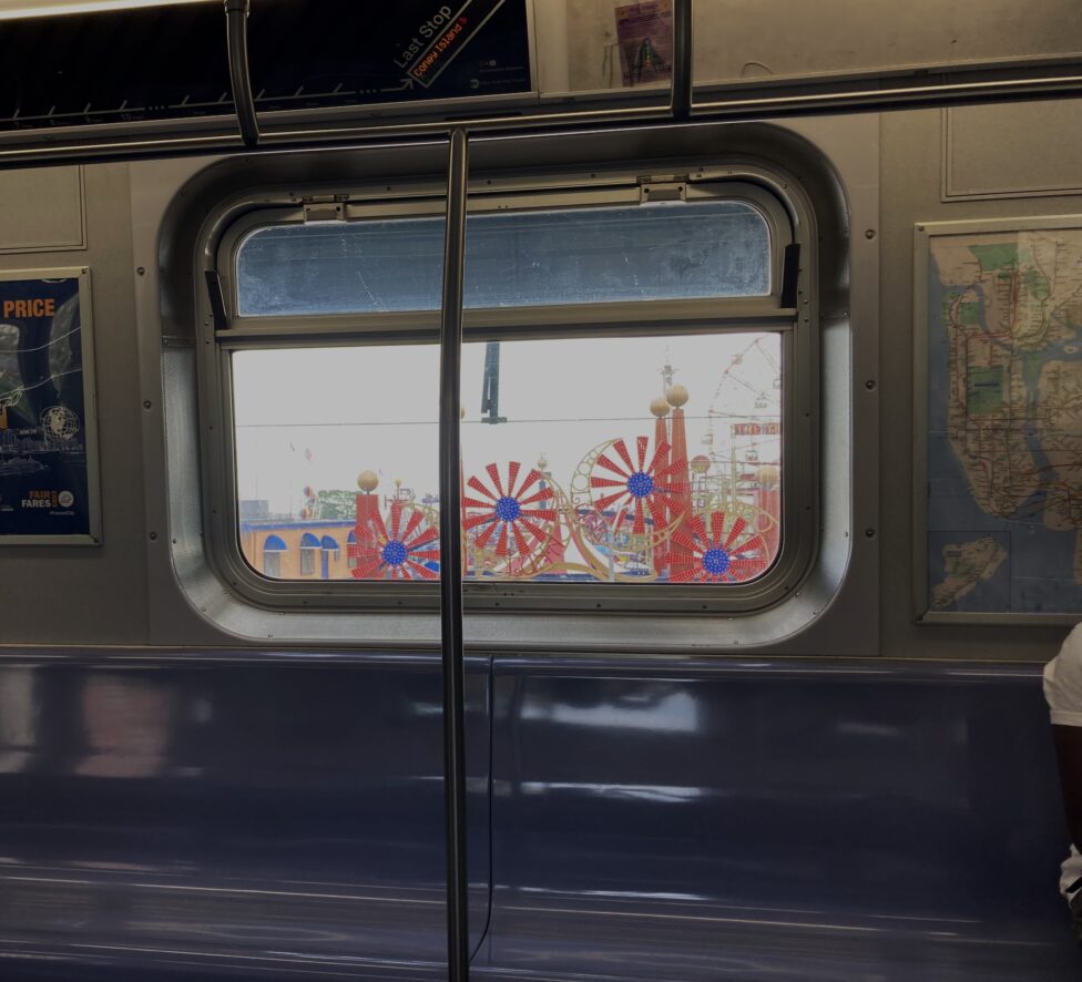 [The interior of a subway cart at its last stop on Coney Island, as indicated on the screen above a window. The window overlooks a colorful amusement park. The bench seating is of a blue color, and a map of New York City hangs to the right of the window.]
