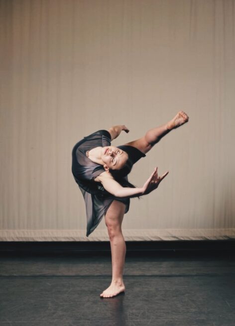 Abigail is wearing a mesh dark multicolor tunic with her dark hair pulled back. She is in motion standing on one foot with the other foot bent and reaching toward her head. She is in front of a white curtain backdrop and her arms are reaching in opposition to each other. Photo was taken by David Rehor