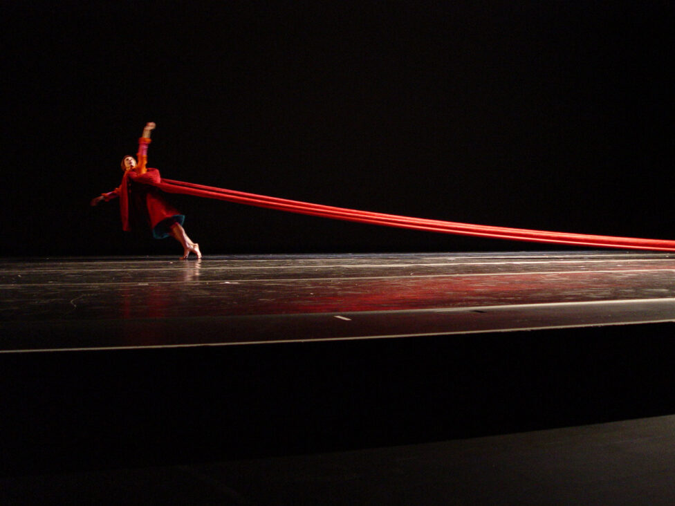 On a darkened stage, dancer Hristoula Harakas wears a red costume whose long train extends behind them. Photo courtesy of the artists.