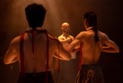 Two dancers turn their backs to the camera while Christopher Unpezverde Núñez looks straight ahead. All three identify the solar plexus in their chests. An amber light bathes them. Photo by Maria Baranova.