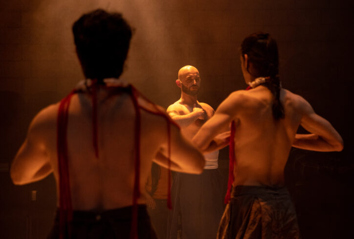 Two dancers turn their backs to the camera while Christopher Unpezverde Núñez looks straight ahead. All three identify the solar plexus in their chests. An amber light bathes them. Photo by Maria Baranova.