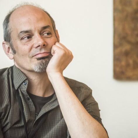 Portrait of Ewan Mclaren. He is looking to the side, Head resting into his propped arm. He wears a light brown button up top over a darker brown t-shirt. Photo courtesy of the artist.