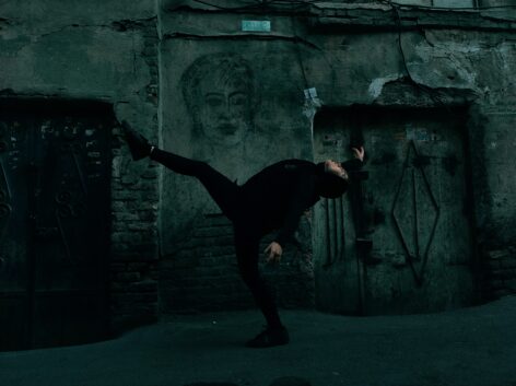 a photo of F.M Sayna who is wearing black. she is dancing in an old downtown place in Tehran bazaar. in the background, you can see a painting on the wall. Photo courtesy of the artist.