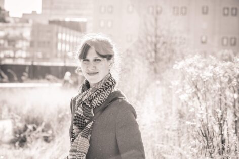 Photo of Anat standing and smiling for a portrait. She is outside wearing a winter jacket and a stripped scarf. In the background there are trees with bare branches. Photo by Elodie Saracco