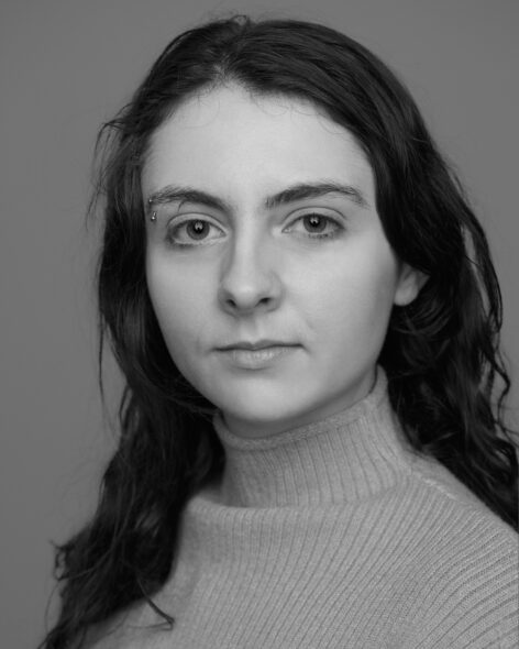 Black and white headshot of Willow, a white non-binary person. Their hair is loose, long and wavy, and they wear a ribbed turtleneck shirt. They gaze into the camera with a serious but gentle expression. Photo by John Helmk