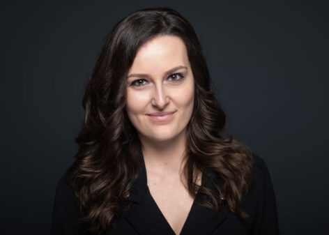 Headshot of Brigitta Kovács. She looks straight into the camera with a grin. She has long dark brown hair and wears a black v neck top. Photo by Dániel Dömölk.