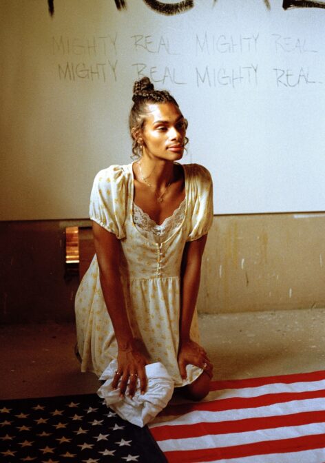 ms. z tye kneels on an American flag with her hands on her lap, while wearing a yellow floral dress. Her hair is up and out of her face. Behind her on a wall are the words mighty real repeated. photo courtesy of artist.