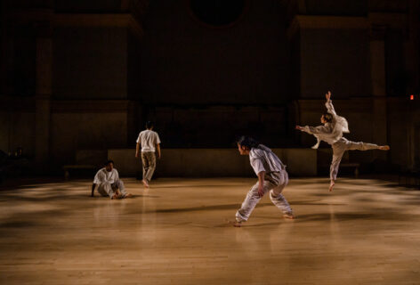Umami Playground Dance Company performs for Movement Research at the Judson Church. They wear white and move around at various levels and facings. Two dancers in motion one standing and the other in a mid squat side stepping. Another dancer crouches to the floor while the last is caught in mid air in a leap. Photo by Rachel Keane.