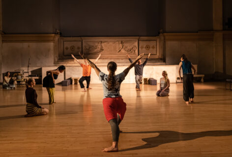 Cayleen Del Rosario performing for movement research at the Judson church. In the foreground a dancer faces away from the camera in a deep lunge with arms extended up and out in straight lines. Two dancers in the background mirror this pose facing towards the camera. on the sides other dancers kneel or stand. Photo by Rachel Keane.