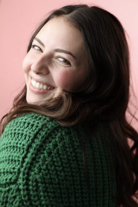 Sophia looks over her shoulder at the camera with a big smile wearing a green sweater and her long brown hair down and curled.