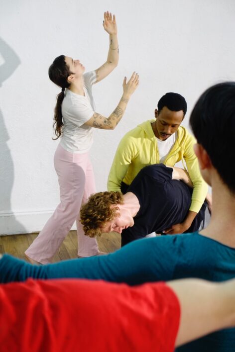 The image takes place in a studio with white walls. Two dancers stand in the foreground, their backs framing a man being held up by another man around the waist upstage. A woman stands beside them looking upward. Photo by Miles Pflanz