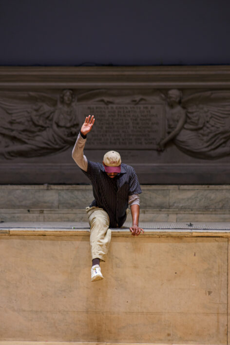 A tall man straddles the edge of a stage in a split. One leg drapes off the front edge and the other is hidden behind him; He holds his right arm in the are and looks down wearing a baseball cap and sneakers. Photo by Rachel Keane.
